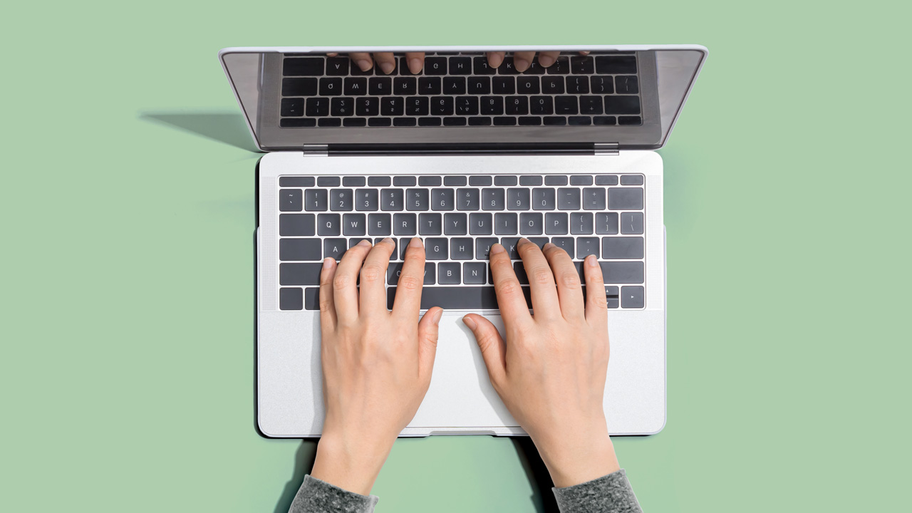 Bilde av en bærbar datamaskin tatt ovenfra. Maskinen står åpen på et bord. På tastaturet ligger to hender.